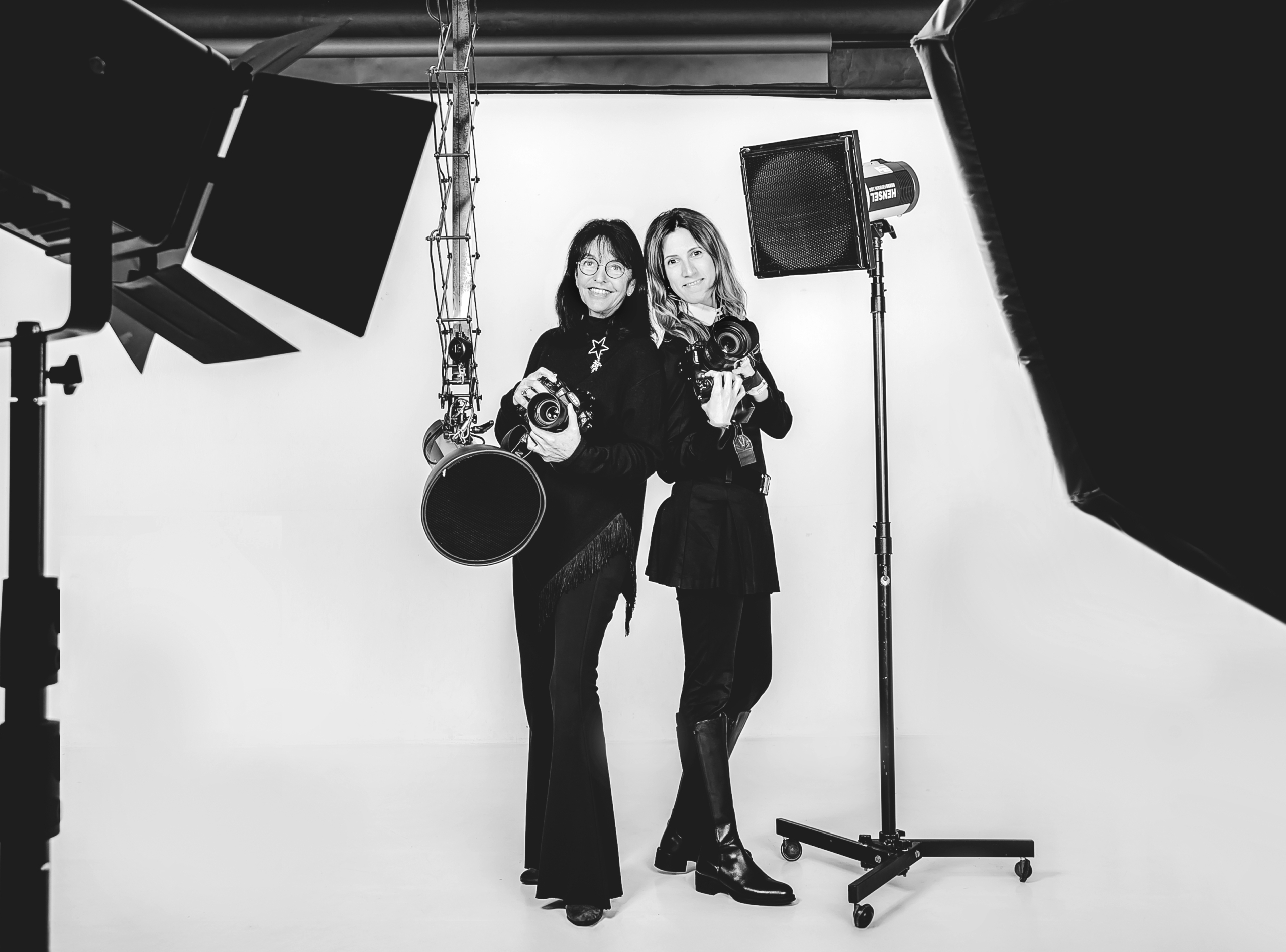 Die Fotografinnen Ilona Voss und Annabell Sibas stehen mit ihren Kameras in den Händen in ihrem Fotostudio in Datteln. Beide lachen in die Kamera und werden von dem professionellen Studio Blitzsystem umrahmt.